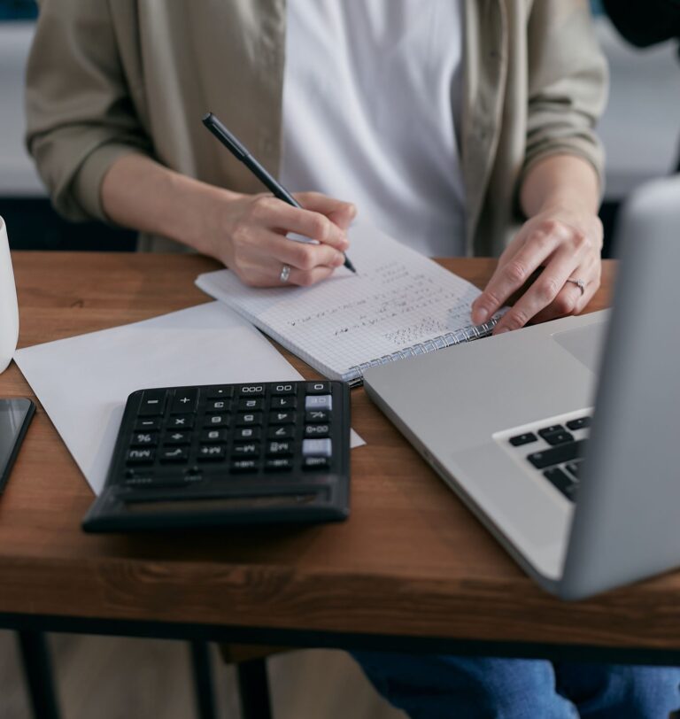Mulher organizando o controle financeiro em caderno, com calculadora e notebook sobre a mesa