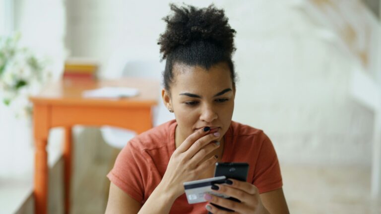 Mulher preocupada com dívidas do cartão confere a fatura no celular com o cartão de crédito na mão